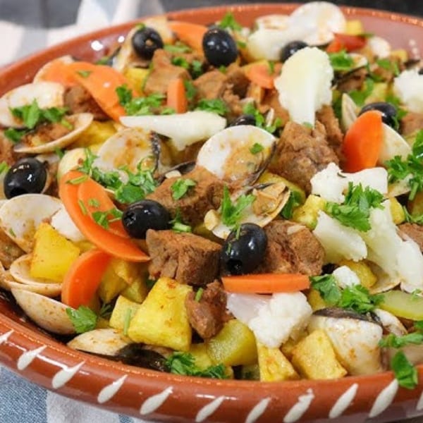 A Portuguese dish with clams, pork, potatoes, carrots, olives, and cauliflower in a terracotta bowl.