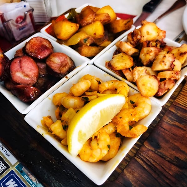 Four white square dishes filled with Portuguese tapas: sausage, potatoes, octopus, and shrimp with lemon.