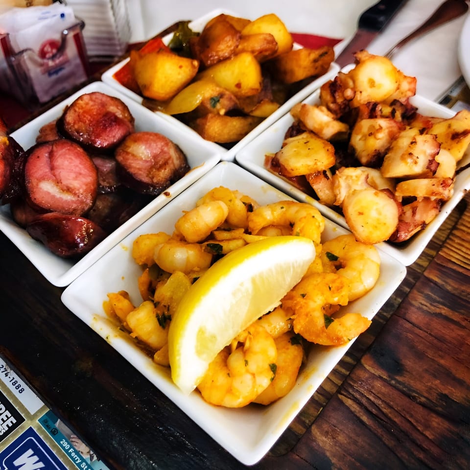 Four white square dishes filled with Portuguese tapas: sausage, potatoes, octopus, and shrimp with lemon.